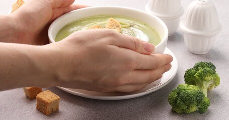 Woman putting bowl of broccoli cream soup onto light grey table, closeup - Powered by Adobe