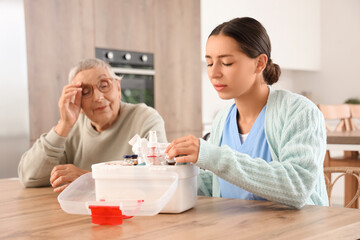 Female caregiver giving pills to senior woman from first aid kit in kitchen at home