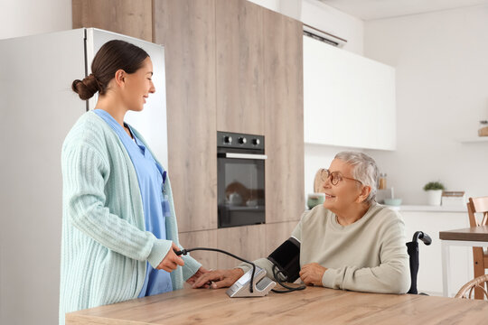 Female caregiver measuring happy senior woman's blood pressure at table in kitchen