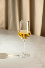 Elegant glass of white wine placed on a marble table with soft curtain background, casting a warm shadow in natural light.