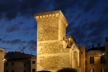 Torre histórica de piedra que se alza en el centro de una plaza al anochecer.  El cielo es de un...