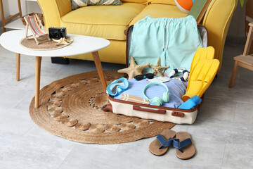 Open suitcase with men's swimwear, headphones and beach accessories on floor near sofa, closeup