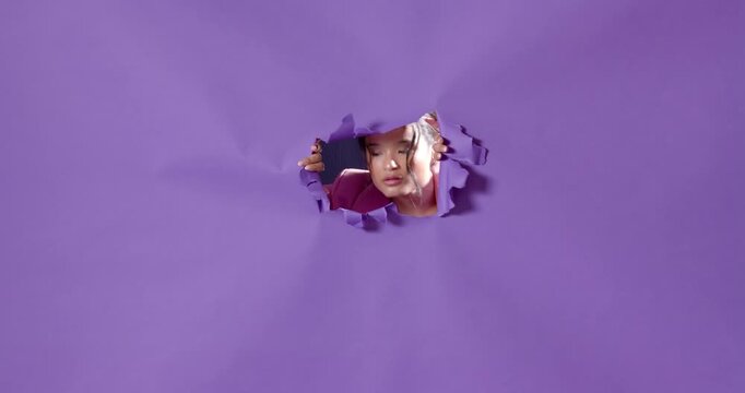 Asian woman in pink blazer breaking through purple paper, looking determined