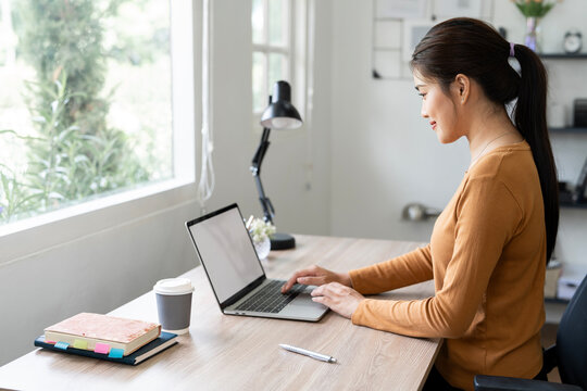 A focused young Asian woman works at her desk on a laptop in a well-lit home office.