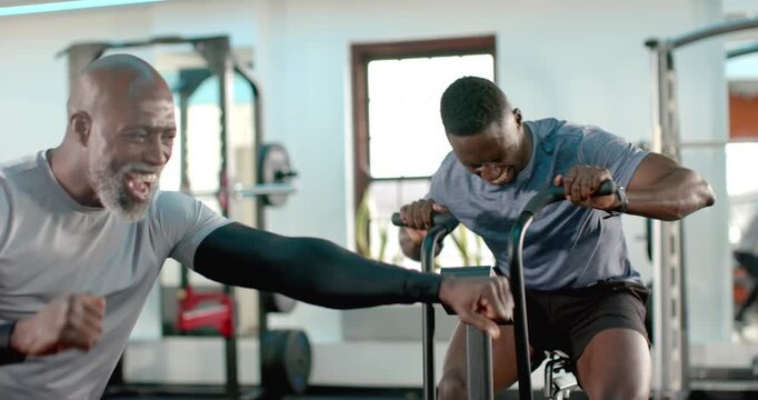 Senior African American trainer punching urging African American man pedaling harder on bike at gym