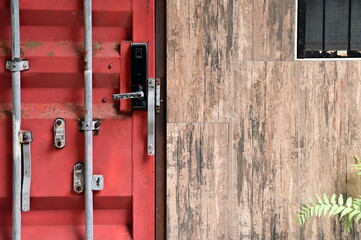 Contrasting door styles featuring red cargo container door with vertical locking bars and wooden textured entrance door with visible handle and decorative plant below.