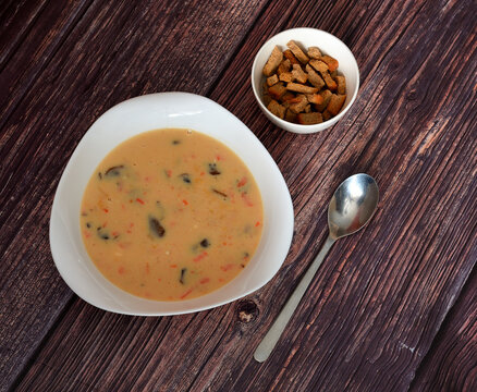 Cream of mushroom and smoked meat soup on a wooden table, a tablespoon and a cup of bread croutons nearby.