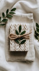 A textured, square soap tied with twine rests on a linen pouch, adorned with green sprigs, set against a softly wrinkled white cloth backdrop