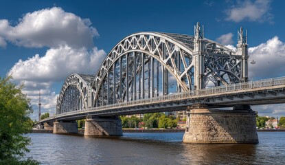 Steel arch bridge spans a wide river on a sunny day with fluffy clouds, anchored by stone supports, creating an impressive cityscape view