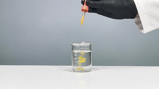 Scientist hand with black glove using dropper to add orange liquid into glass beaker, laboratory experiment on gray background