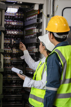 teamwork in industrial maintenance, workplace safety, and modern energy technology, Engineers wearing safety helmets and reflective vests inspecting electrical control panel with tools and checklist
