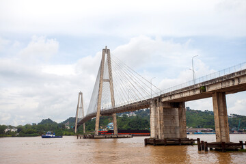 The magnificent Mahkota II Bridge spanning the Mahakam River in Samarinda, Borneo. A tugboat pulls a coal barge underneath, showcasing the river's vital role for industrial transport.