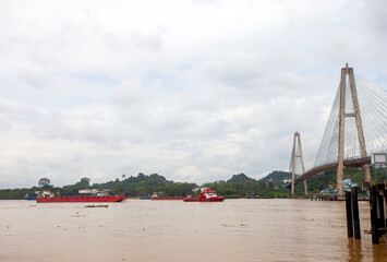 The magnificent Mahkota II Bridge spanning the Mahakam River in Samarinda, Borneo. A tugboat pulls a coal barge underneath, showcasing the river's vital role for industrial transport.