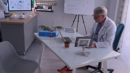 Male doctor in white coat and glasses studies patient information on a digital tablet at a desk with laptop and medical charts. Concept of telemedicine, health records and modern medical technology. - Powered by Adobe