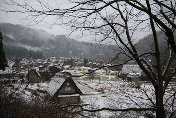 A panoramic view of the historic Shirakawa-go village, with farmhouses and rice fields covered in the season's first snow on a gloomy and misty day.