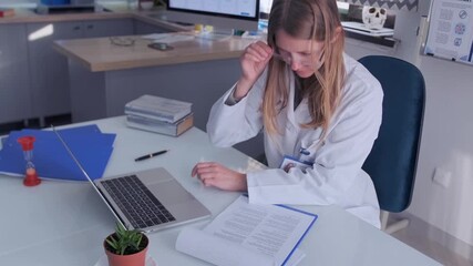 Female doctor in white coat uses laptop at desk with books and folders in hospital office. Scene of medical professional analyzing healthcare data and patient information.  - Powered by Adobe