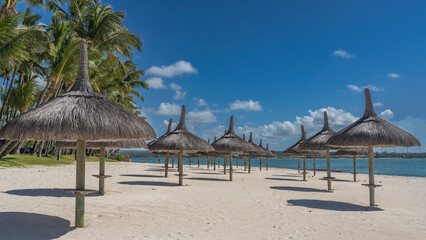 Rows of straw umbrellas on a sandy beach without people. Round shadows on white sand. Turquoise ocean. Palm trees on a background of blue sky, clouds. Mauritius. Resort. Hotel. Low season.