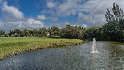 Golf course. Holes, fairway bunkers, flags on the green lawn. The fountain jet in the lake. Circles on the water. Tropical vegetation, palm trees in the distance. Blue sky, clouds. Resort. Hotel.