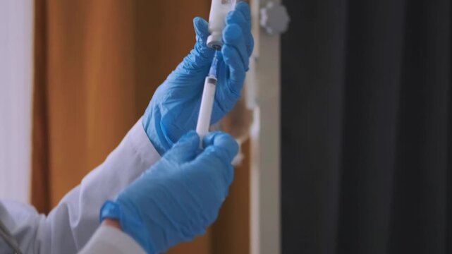 Female doctor in white coat and blue gloves drawing medicine into syringe from vial next to hospital bed with patient on IV drip, preparing injection and treatment procedure in clinic.