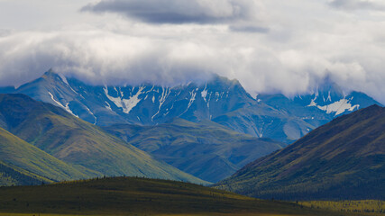 A panoramic view of a mountain range under a cloudy sky. Snow-capped peaks rise in the background, partially hidden by clouds, while lush green and brown hills fill the foreground.