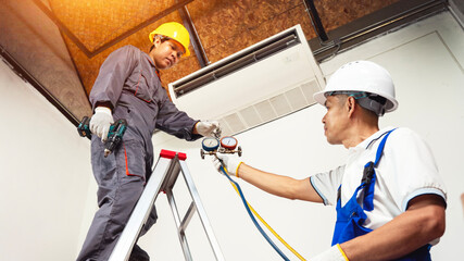 HVAC installation: Two professional technicians, in their safety helmets and workwear, diligently install a new air conditioning unit indoors.
