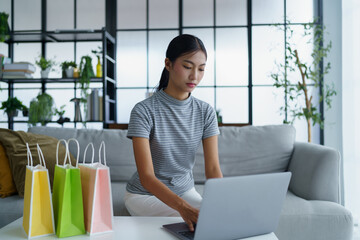 Asian woman sitting on sofa with shopping bags on table her using laptop shopping online in living room at home