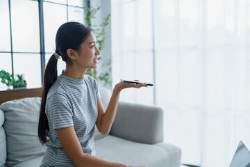 Asian woman sitting on sofa and holding smartphone near mouth talking with AI. using voice assistance at home