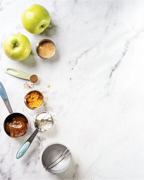 Food background with baking ingredients &mdash; apples, sugar, caramel, starch, cardamom, and orange zest on marble surface, left side layout with copy space for text.