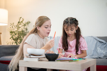 Caucasian mother holding color pen talking with daughter drawing at table aside sofa in living room.