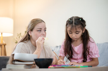 Caucasian mother eating snack beside kid daughter doing homework at table aside sofa in living room.