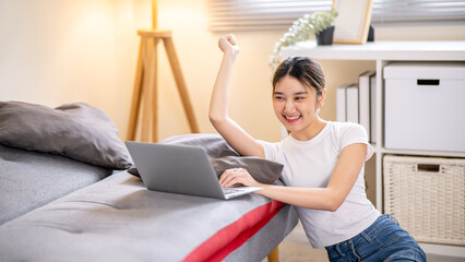 Happy asian woman raising a hand looking at laptop while sitting leaning against sofa bed on floor.