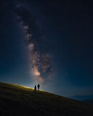 Fototapeta premium Pareja contemplando la Vía Láctea en una colina iluminada de noche, paisaje estelar romántico y majestuoso