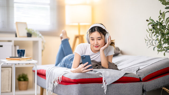 Asian woman putting head on hand looking at phone while listening to music in headphones on sofa bed