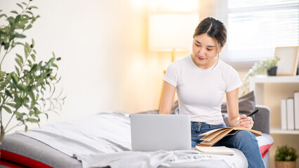 Asian woman holding pen and notebook on lap looking at laptop and sitting on sofa bed in living room
