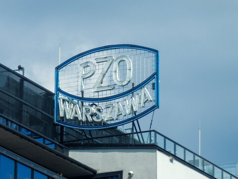 A glowing PZO Warszawa sign on the rooftop of a modern building, captured at dusk against a cloudy sky.