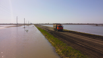 Fototapeta premium An orange dump truck drives down a narrow dirt road, surrounded by a vast flooded landscape, symbolizing the challenging conditions faced in remote areas
