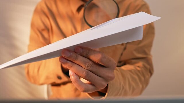A person uses a magnifying glass to examine a paper rocket in an office with a laptop, symbolizing cautious adoption of innovation and the importance of evaluating new ideas before launching them.
