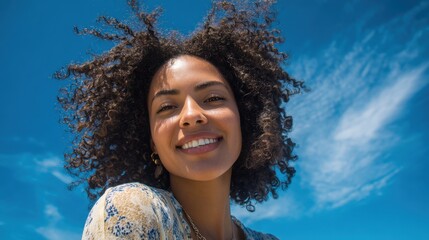 A woman with beautiful curly hair enjoys a sunny day at the beach. She smiles widely, with the clear blue sky and soft clouds providing a perfect backdrop for her joyful expression.