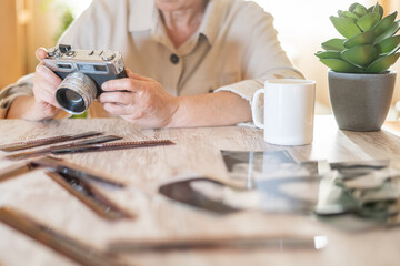 Senior woman at a table holding a classic film camera and looking at old film negatives and prints,...