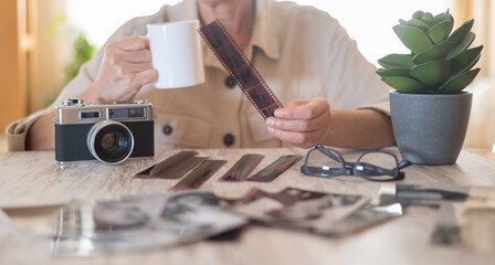 Senior woman at home holding a film strip, reviewing old memories from analog photography on a wooden table with a vintage camera, coffee cup, developed prints, and eyeglasses