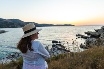 Woman in a sun hat stands on a rocky cliffside, arms crossed, gazing at the serene sea during golden hour