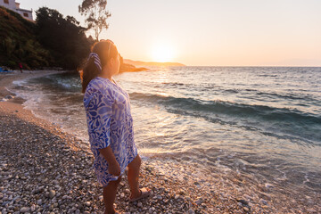 Woman in Patterned Dress Standing on Pebbled Beach at Sunset, Gazing at Ocean