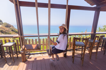 Smiling girl in a sun hat sits on a wooden swing, looking back at the camera with a scenic ocean view behind her