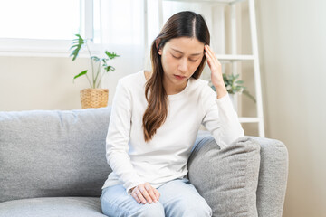 Dizzy asian young woman, girl headache or migraine pain suffering from vertigo while sitting on couch in living room at home, holding head with hand, health problem of brain or inner ear not balance.