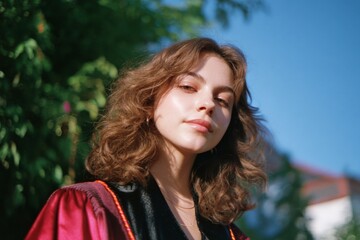 Young caucasian female graduate in red gown posing outdoors on a sunny day