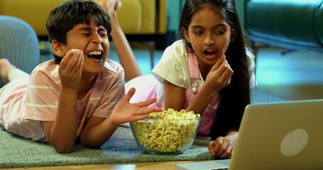 Indian Asian small sibling kids watching funny serial on laptop while eating popcorn, sitting on carpet in modern living room, enjoying entertainment, leisure activity, bonding, happy childhood - Powered by Adobe