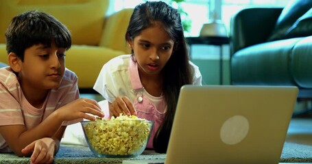 Indian Asian small sibling kids watching funny serial on laptop while eating popcorn, sitting on carpet in modern living room, enjoying entertainment, leisure activity, bonding, happy childhood - Powered by Adobe