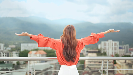Rear view of woman raising arms with confidence while standing on rooftop overlooking city and mountains