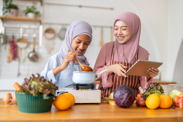Muslim girl cooking with woman using tablet for recipe, halal food learning with technology, warm islamic family lifestyle, digital nutrition education for muslim kid, modern halal kitchen inspiration