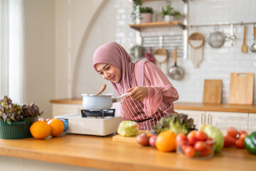 Muslim woman happy cooking healthy meal in kitchen wearing hijab making soup in pot with fresh vegetables food preparation activity nutrition, ramadan healthy recipe preparation, islamic food culture